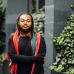 Immanuel Wilkins met baard en goudkleurige bril, zwart shirt en rode sjaal met groene streep over de schouders, armen gekruist tussen pilaren met klimop.