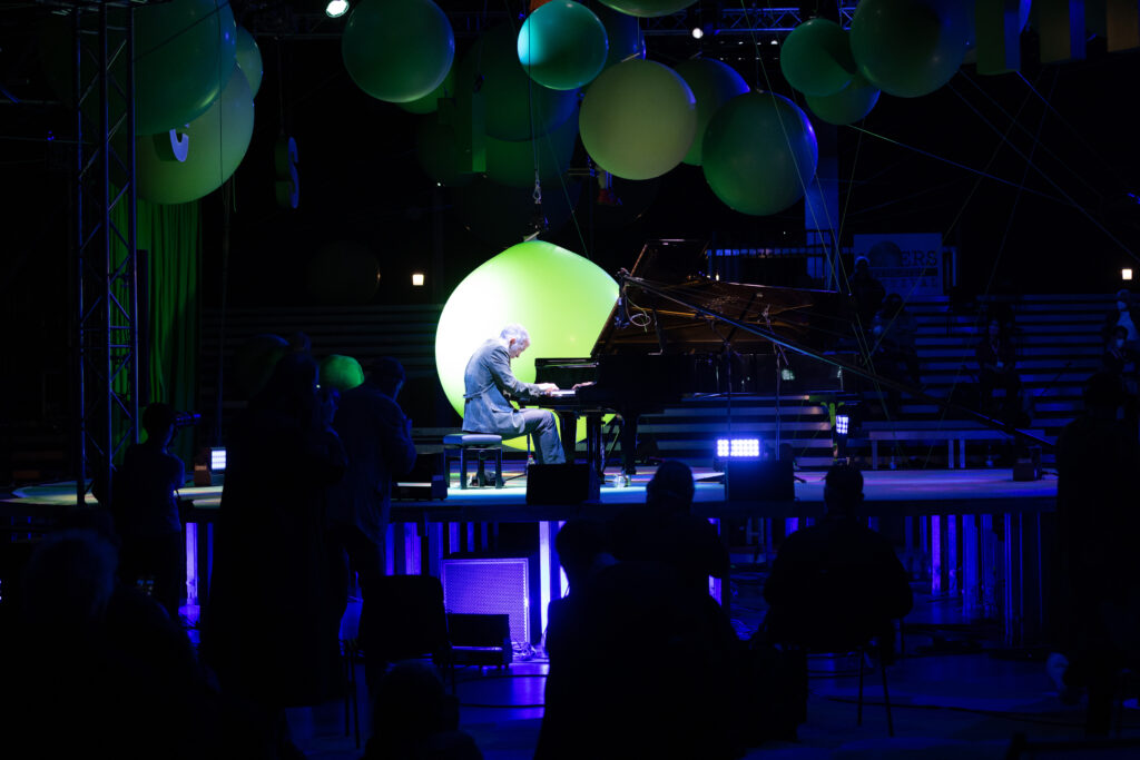 Pianist aan een vleugel op een podium met grote groene ballonnen, het publiek op de voorgrond als silhouet