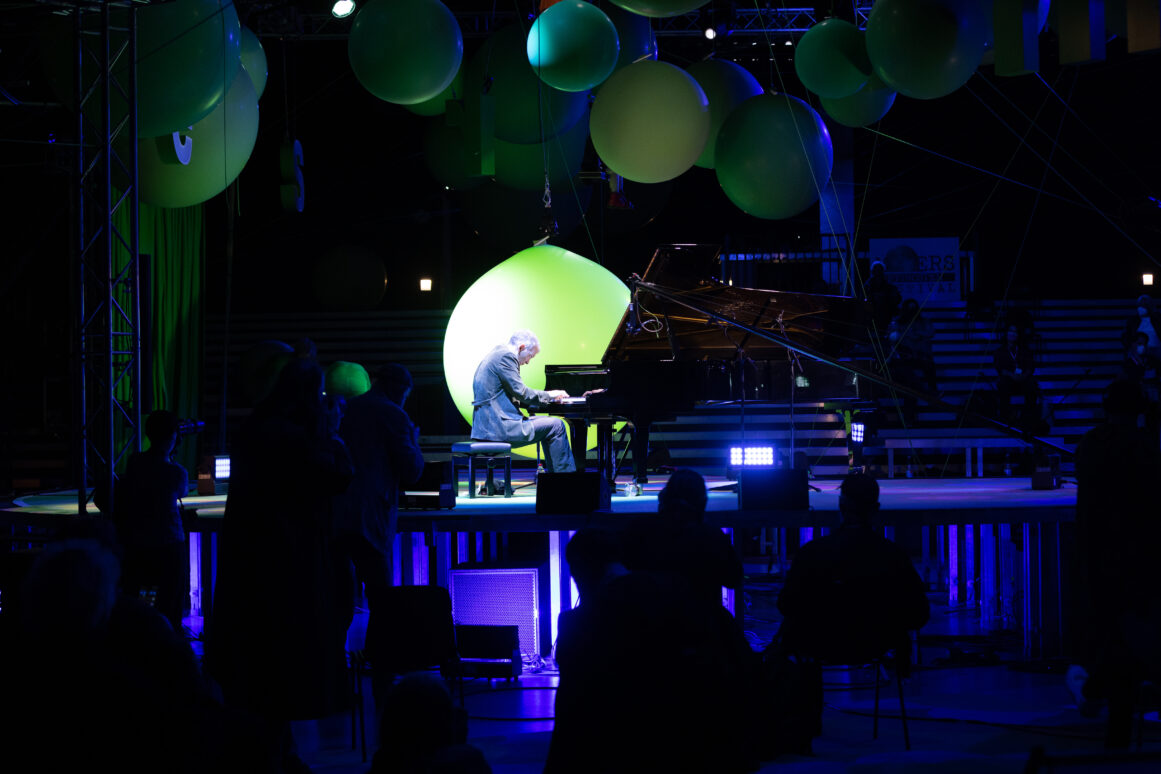 Pianist aan een vleugel op een podium met grote groene ballonnen, het publiek op de voorgrond als silhouet