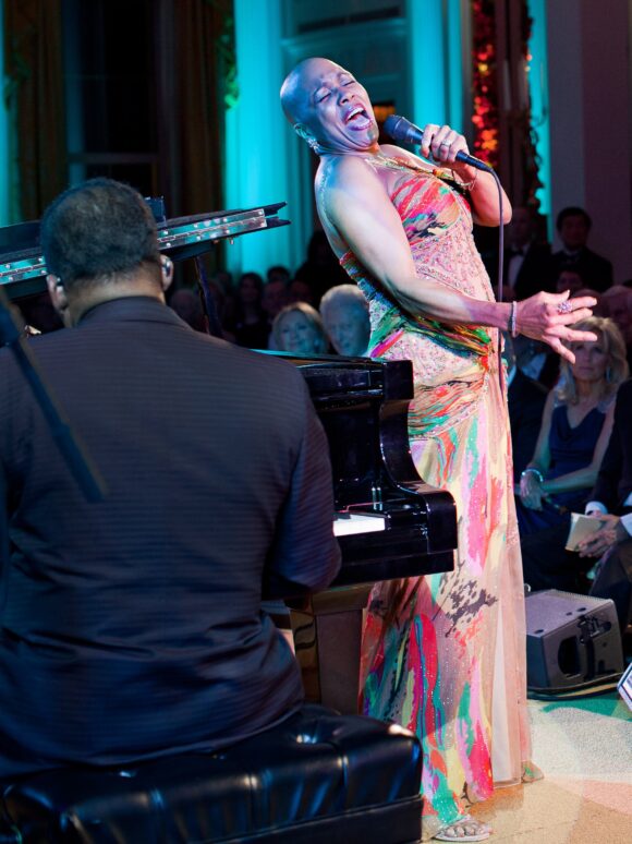 Dee Dee Bridgewater zingt met de microfoon in de hand en het hoofd lachend achterover, in een veelkleurige jurk, naast een vleugel in het Witte Huis (2011).