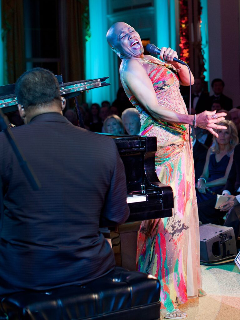 Dee Dee Bridgewater zingt met de microfoon in de hand en het hoofd lachend achterover, in een veelkleurige jurk, naast een vleugel in het Witte Huis (2011).