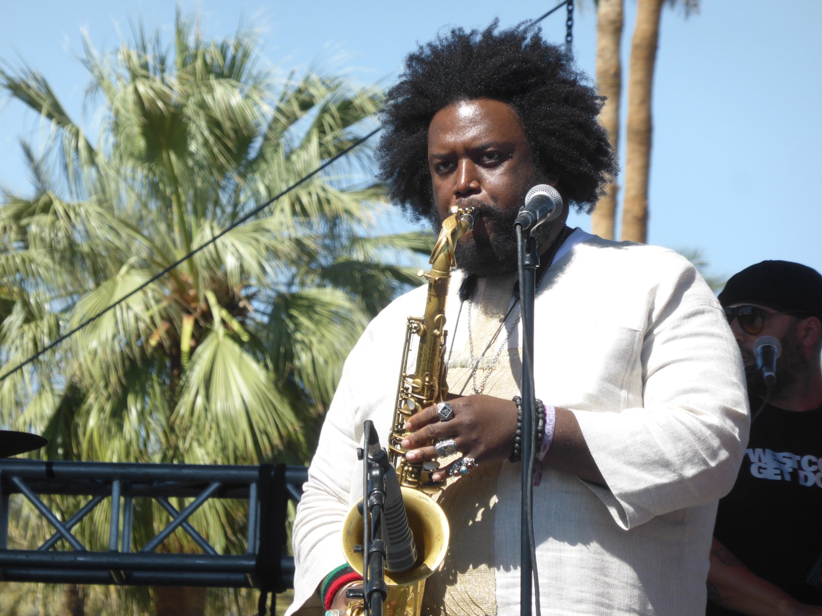 Saxofonist met baard en afrokapsel, in een wit overhemd, achter een microfoon op een podium buiten, met palmbomen op de achtergrond.