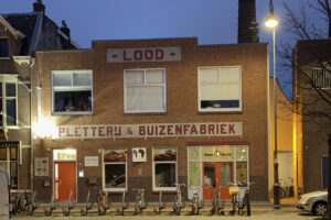 Avondopname van de bakstenen gevel van De Pletterij aan de Lange Herenvest in Haarlem, met de opschriften Lood en Pletterij & Buizenfabriek, fietsen voor de deur en een fabrieksschoorsteen erachter.
