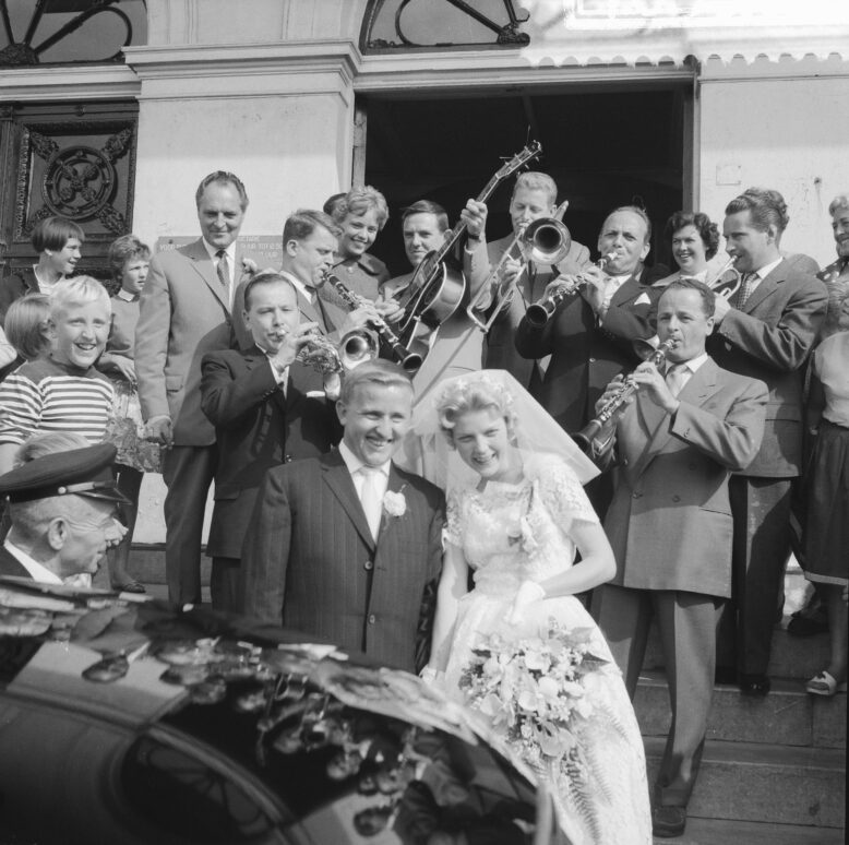 Een bruidspaar op de trap voor een ingang, omringd door musici met trompetten, trombones en een gitaar, met toeschouwers op de voorgrond; zwart-witfoto uit 1959.